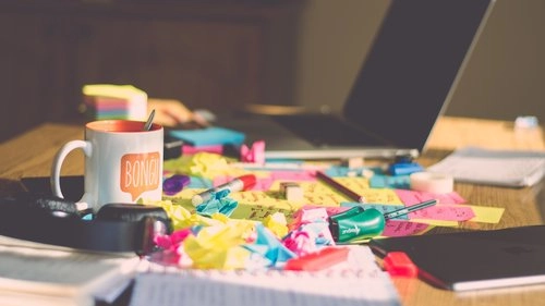 A cluttered home office desk with sticky notes, a coffee mug, and a laptop, illustrating ideas for home office organization and workspace decluttering tips by dClutterfly.com