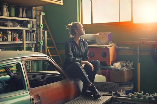 Person sitting in a cluttered garage, gazing thoughtfully toward the sunlight streaming through the window, symbolizing emotional blocks associated with decluttering.