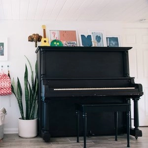 Black upright piano in a bright and organized room, surrounded by potted plants, artwork, and hanging storage bags for a tidy aesthetic.