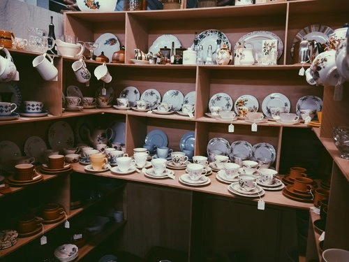 A well-organized display of vintage and antique dinnerware, including plates, teacups, and mugs, in a wooden shelving unit at an appraisal or auction house.