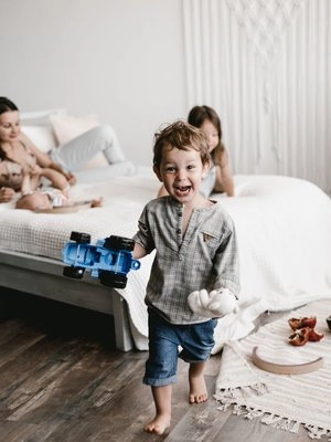 A playful young boy holding a toy truck and stuffed animal, running joyfully in a bedroom while other children sit on a neatly made bed in the background.